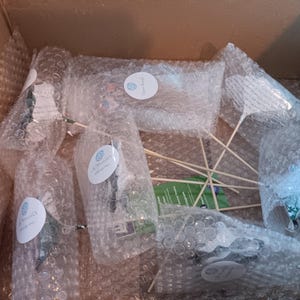 May include: A cardboard box containing several items wrapped in bubble wrap. The items, possibly decorations for an event, are secured with wooden sticks. Each item is sealed with a round sticker, suggesting a branded product.