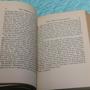 May include: Open book with text on aged paper. The left page displays the title "THE ARABIAN NIGHTS" and the right page reads "THE STORY OF ALNASCHAR". The book is held open by a hand.