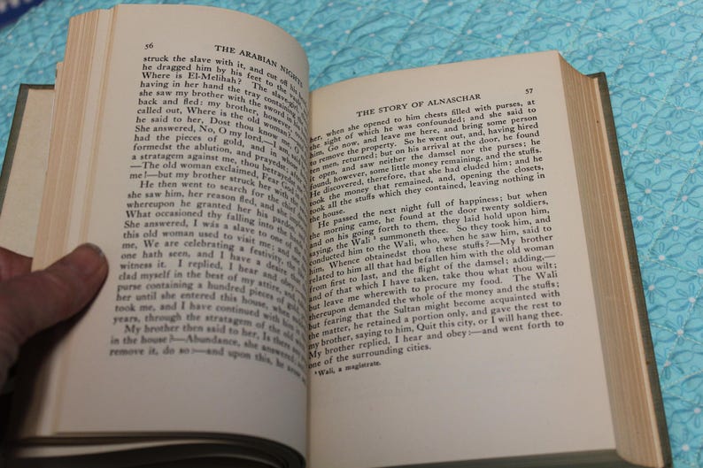 May include: Open book with text on aged paper. The left page displays the title "THE ARABIAN NIGHTS" and the right page reads "THE STORY OF ALNASCHAR". The book is held open by a hand.