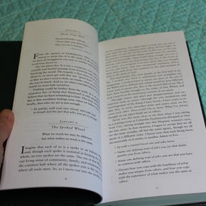 Może przedstawiać: Otwarta książka z czarnymi okładkami, trzymana w dłoni. Strony są wypełnione tekstem. Lewa strona zawiera tytuł "The Spoked Wheel" i tekst "What we reach for may be different, but what makes us reach is the same."