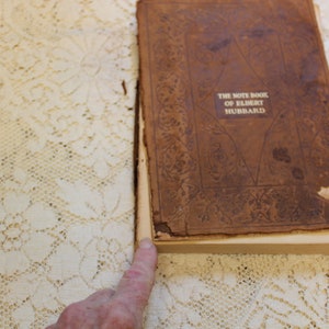 May include: The front cover of a brown leather-bound book with gold lettering that reads "The Note Book of Elbert Hubbard".