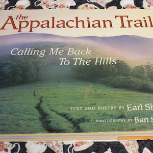 May include: A hardcover book titled "The Appalachian Trail" with the subtitle "Calling Me Back To The Hills." The cover features a landscape of green fields, a winding path, and distant mountains. Text and poetry by Earl Shaffer, photography by Bart Smith.