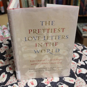 May include: A book titled "The Prettiest Love Letters in the World" with text in blue and red. The book is standing upright on a floral patterned surface. The book's title is the main focus.