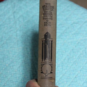 May include: A vintage book titled "The Arabian Nights" with text in black on a tan cover. The spine features illustrations and the names of the editor and illustrator. The book is held by a hand.