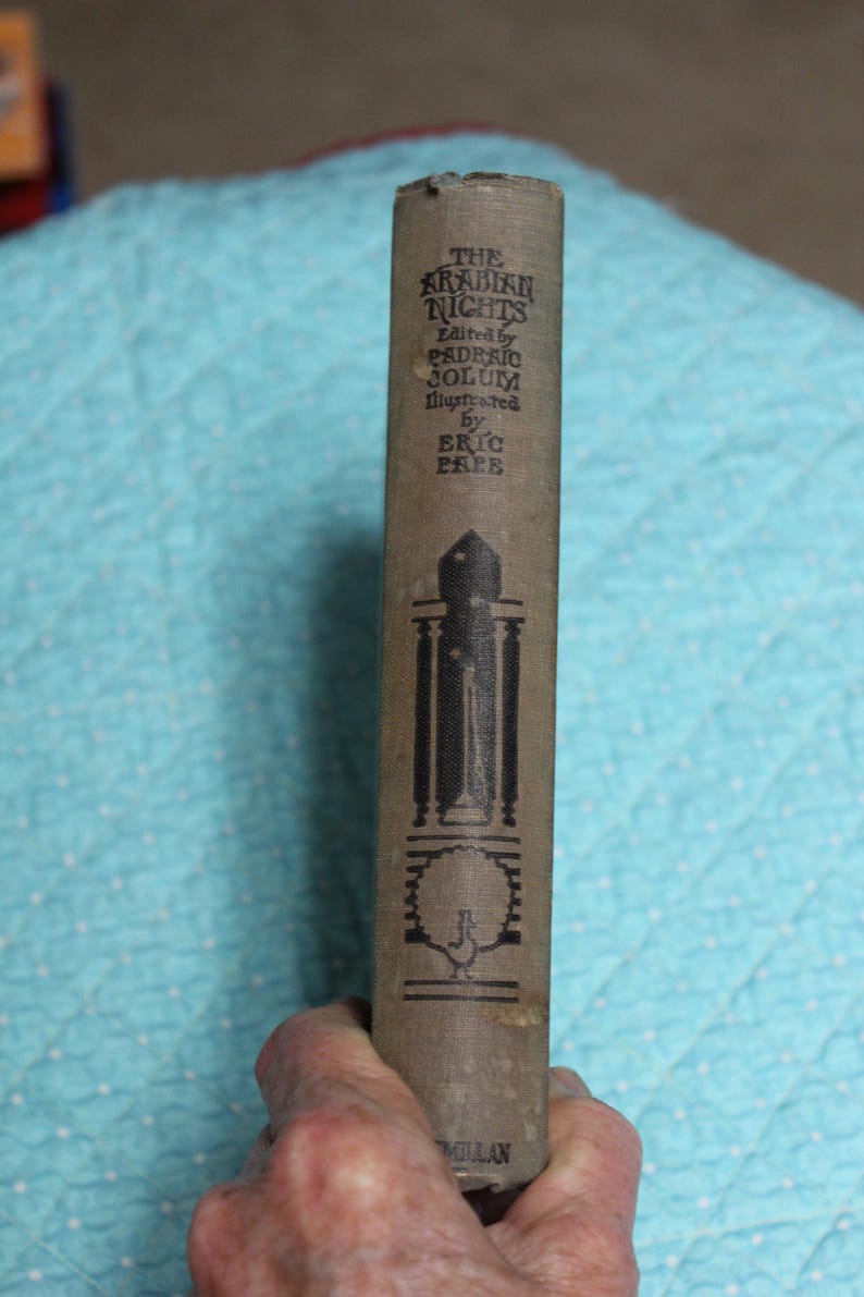 May include: A vintage book titled "The Arabian Nights" with text in black on a tan cover. The spine features illustrations and the names of the editor and illustrator. The book is held by a hand.