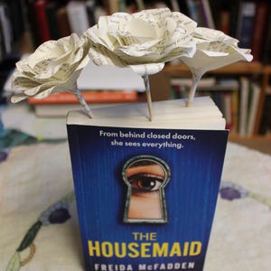 May include: A copy of "The Housemaid" by Freida McFadden, with a blue cover and a keyhole design. Three paper roses, crafted from book pages, are arranged on top of the book, set against a backdrop of bookshelves.