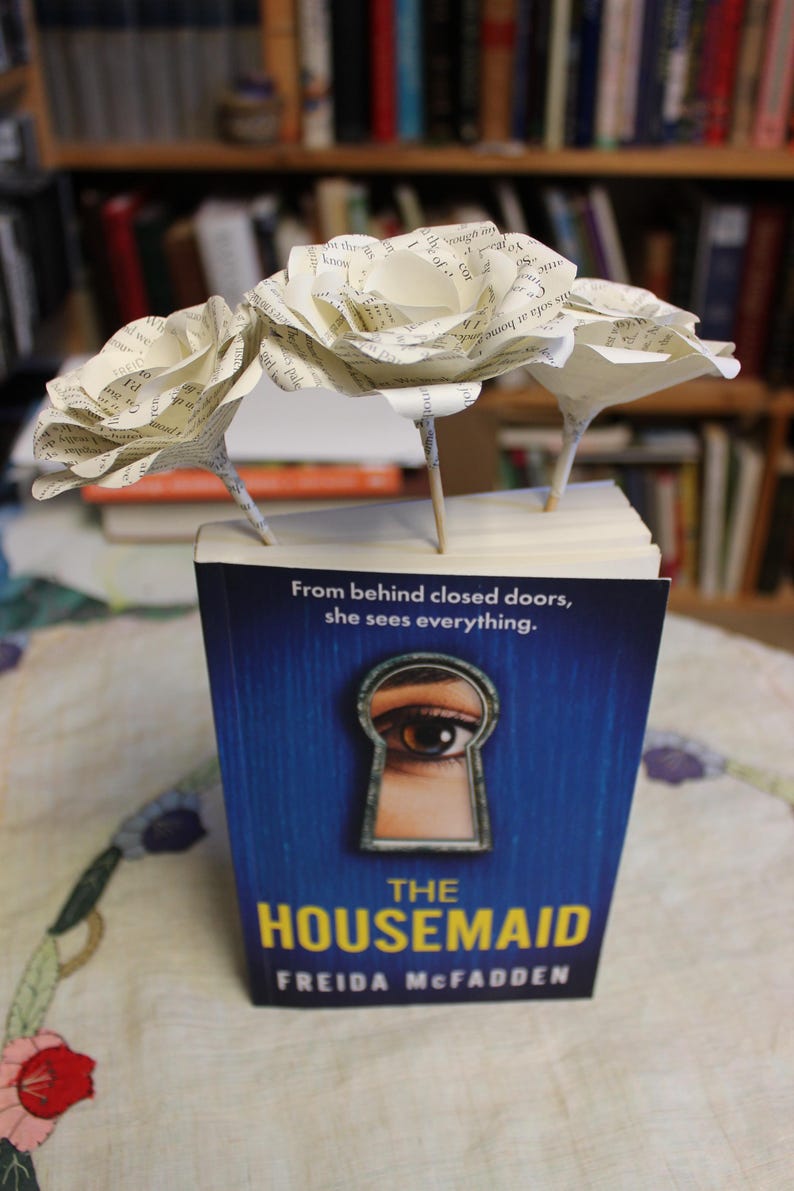 May include: A copy of "The Housemaid" by Freida McFadden, with a blue cover and a keyhole design. Three paper roses, crafted from book pages, are arranged on top of the book, set against a backdrop of bookshelves.