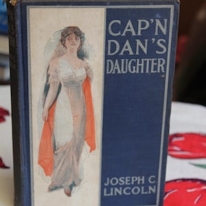 May include: A vintage book cover with a blue background and the title "Cap'n Dan's Daughter" in white lettering. The cover features an illustration of a woman in a white dress and a red shawl.