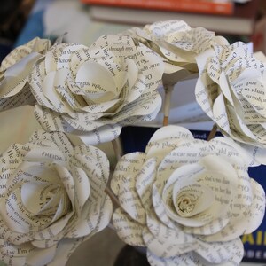 May include: A bouquet of paper roses made from book pages. The white flowers, with visible text, are arranged in a circular shape. The stems are visible, creating a delicate and artistic display. A unique floral arrangement.