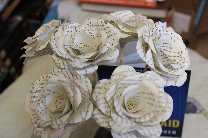 May include: A bouquet of paper roses made from book pages. The white flowers, with visible text, are arranged in a circular shape. The stems are visible, creating a delicate and artistic display. A unique floral arrangement.
