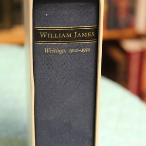 May include: A blue hardcover book with a white slipcase. The spine features a black rectangular label with gold lettering that reads William James, followed by the text Writings, 1902-1910 in a smaller gold font below the label.
