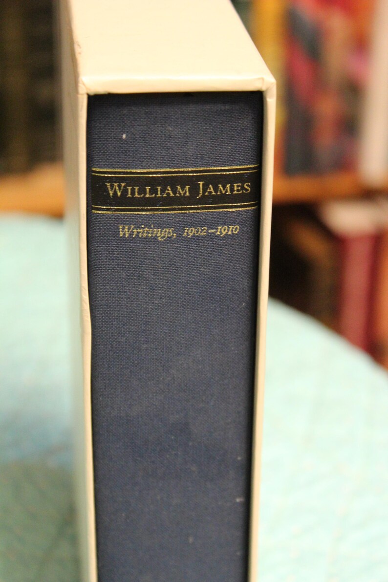 May include: A blue hardcover book with a white slipcase. The spine features a black rectangular label with gold lettering that reads William James, followed by the text Writings, 1902-1910 in a smaller gold font below the label.