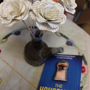 May include: A display of paper flowers crafted from book pages in a small vase, alongside a book titled "The Housemaid" by Freida McFadden. The book's cover is blue with a keyhole illustration.