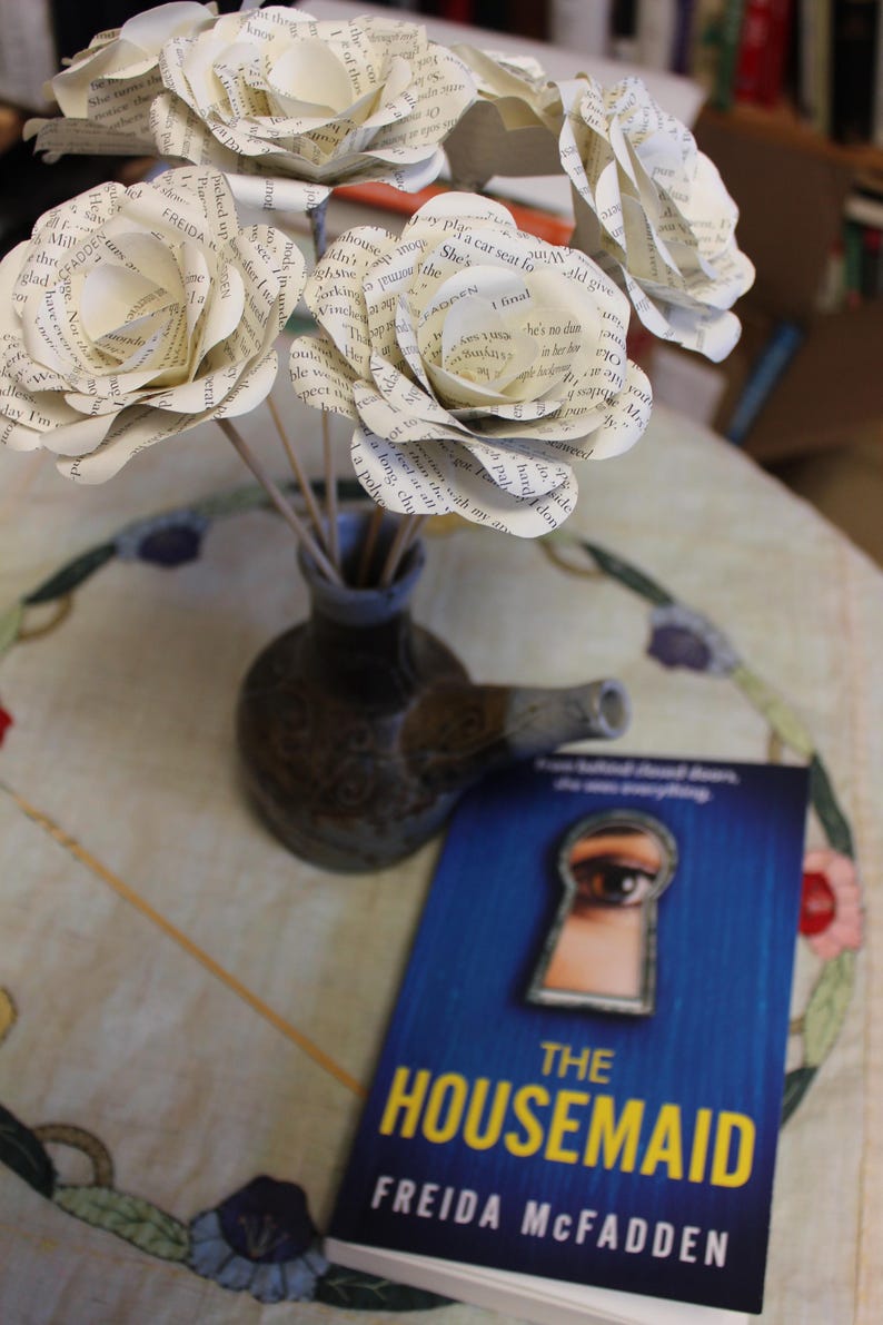 May include: A display of paper flowers crafted from book pages in a small vase, alongside a book titled "The Housemaid" by Freida McFadden. The book's cover is blue with a keyhole illustration.