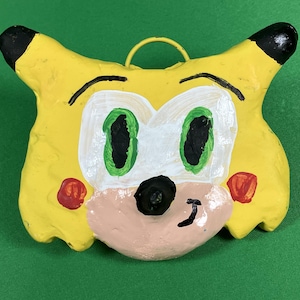 May include: A yellow, hand-painted ceramic ornament shaped like a cartoon character with green eyes, a black nose, and red cheeks. The ornament has a metal loop for hanging.