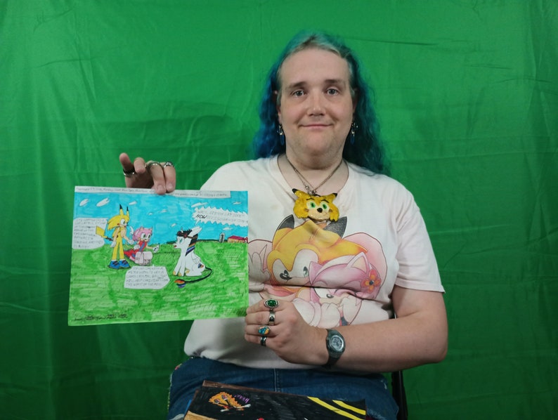 May include: A person holding a colorful drawing featuring cartoon characters in a grassy field. The drawing includes speech bubbles with text. The person is wearing a light-colored t-shirt with a cartoon character design and a watch.
