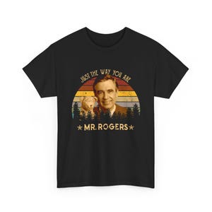 May include: A black short-sleeve t-shirt featuring a graphic of Mr. Rogers and a small puppet. The design includes a sunset background with horizontal stripes, a forest silhouette, and the text "JUST THE WAY YOU ARE" and "MR. ROGERS".