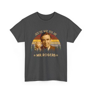 May include: A dark gray short-sleeve t-shirt featuring a graphic of Mr. Rogers and a small dog. The design includes a retro-style sunset background with pine trees, the text "JUST THE WAY YOU ARE" at the top, and "MR. ROGERS" with stars below.