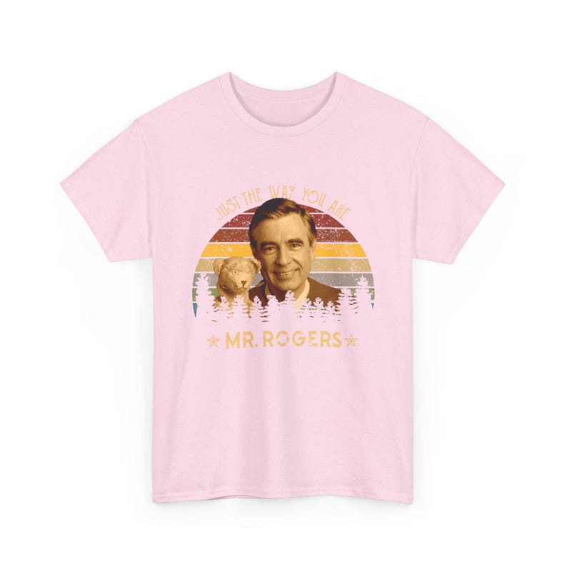 May include: A light pink short-sleeve t-shirt featuring a graphic of Mr. Rogers and a small dog. The design includes a sunset background with horizontal stripes, a forest silhouette, and the text "JUST THE WAY YOU ARE" and "MR. ROGERS".