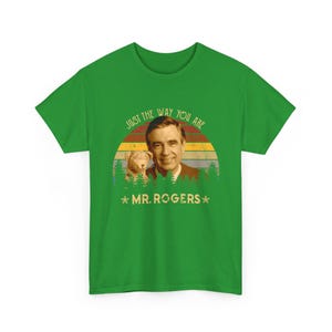 May include: A bright green short-sleeve t-shirt featuring a graphic of Mr. Rogers and a puppet. The design includes a sunset background with pine trees and the text "JUST THE WAY YOU ARE" and "MR. ROGERS" printed in a retro-style font.