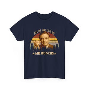 May include: A navy blue t-shirt featuring a graphic print of Mr. Rogers and a small lion puppet. The design includes a sunset background with horizontal stripes, a forest silhouette, and the text "JUST THE WAY YOU ARE" and "MR. ROGERS".