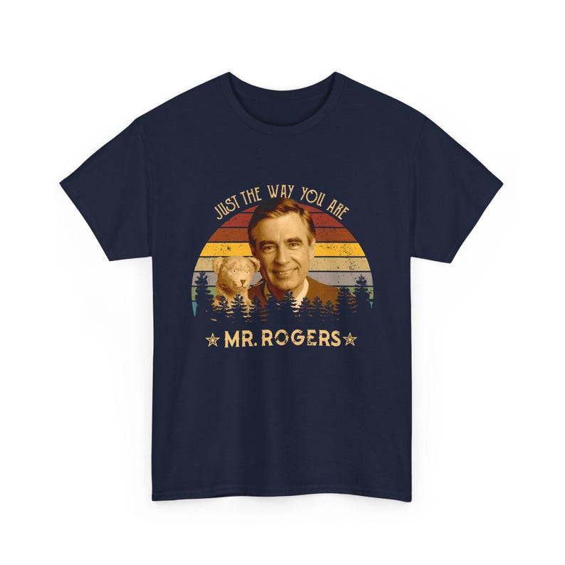 May include: A navy blue t-shirt featuring a graphic print of Mr. Rogers and a small lion puppet. The design includes a sunset background with horizontal stripes, a forest silhouette, and the text "JUST THE WAY YOU ARE" and "MR. ROGERS".