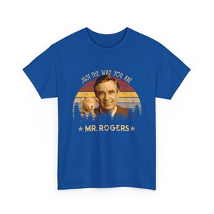 May include: A royal blue t-shirt featuring a graphic print of Mr. Rogers and a small dog. The design includes a sunset background with horizontal stripes, a forest silhouette, and the text "JUST THE WAY YOU ARE" and "MR. ROGERS" with stars.