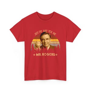 May include: A red short-sleeved t-shirt featuring a graphic of Mr. Rogers and a puppet. The design includes a sunset background with horizontal stripes, a forest silhouette, and the text "JUST THE WAY YOU ARE" and "MR. ROGERS" with two stars.