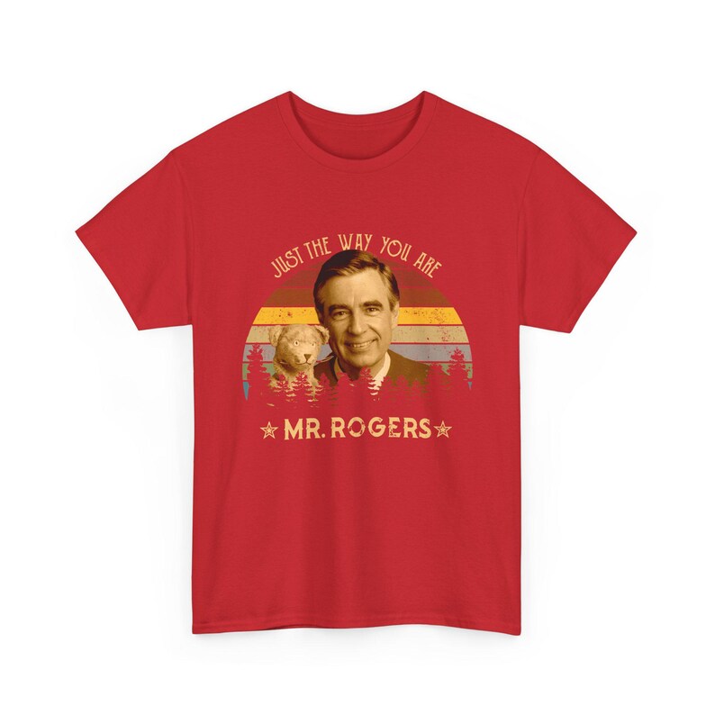 May include: A red short-sleeved t-shirt featuring a graphic of Mr. Rogers and a puppet. The design includes a sunset background with horizontal stripes, a forest silhouette, and the text "JUST THE WAY YOU ARE" and "MR. ROGERS" with two stars.