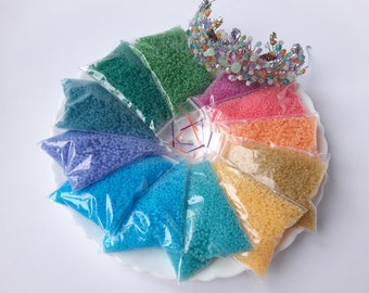 May include: Assortment of small seed beads in various colours, packaged in clear plastic bags. The beads are arranged in a circle on a white plate, with a silver and colourful crown in the centre.