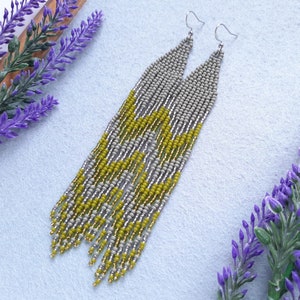 May include: A pair of long, beaded earrings with a geometric pattern in shades of gray, yellow, and gold. The earrings are made with small, seed beads and have a fringe at the bottom.