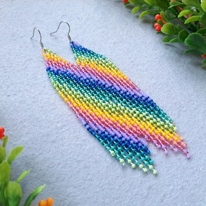 Puede incluir: Pendientes de cuentas de arcoíris con ganchos plateados. Los pendientes son largos y presentan un degradado de colores, incluyendo rojo, naranja, amarillo, verde, azul, añil y violeta.