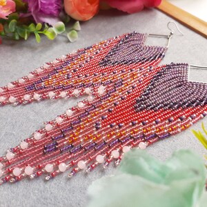 May include: A pair of long, beaded earrings with a geometric pattern in shades of pink, purple, and orange. The earrings are made with small seed beads and feature larger pink beads at the bottom.