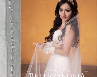 May include: A bride wearing a white wedding dress and a veil with a beaded edge. She is holding the veil in her hands and looking at the camera. The background is a light orange wall. The text "BELLEZANOVA" is visible in the lower center of the image.