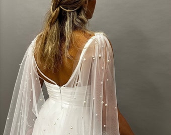 May include: A white wedding dress with a sheer, pearl-studded cape. The dress features a low back and spaghetti straps. The cape drapes over the shoulders and is adorned with scattered pearls, creating an elegant and romantic look.