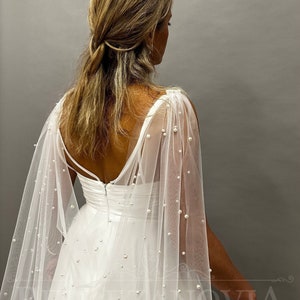 May include: A white wedding dress with a sheer, pearl-studded cape. The dress has a low back and spaghetti straps. The cape drapes over the shoulders and is decorated with scattered pearls, creating an elegant and romantic look.