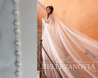 May include: A long, white bridal veil with a beaded edge. The veil is draped over a staircase and the bride is standing on the stairs. The text "BELLEZANOVA" is visible at the bottom of the image.