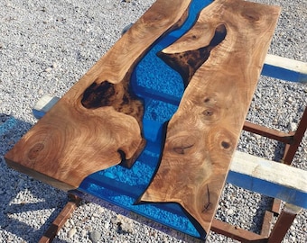 May include: A rectangular wooden table with a river of blue resin running through the center. The wood has a natural grain and knot pattern. The table is supported by a metal frame.