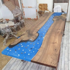 May include: A handcrafted wooden table with a river of blue epoxy resin running through the center. The resin is embedded with small, colorful stones. The table has a natural wood grain finish and is supported by a metal frame.