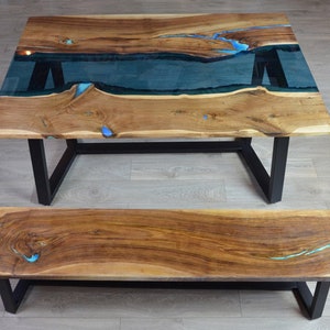 May include: A wooden dining table and bench set. The table features a live edge design with a clear epoxy river and blue accents. The table and bench have black metal legs. The wood has a natural grain and warm tones.