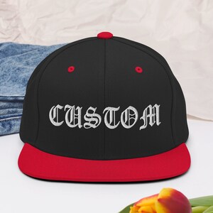 Può includere: Un cappello da baseball nero e rosso con la parola "CUSTOM" ricamata in bianco in un font inglese antico.