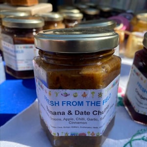 Pode incluir: Um frasco de vidro de chutney de banana e tâmara com uma tampa dourada. O rótulo diz "Fresh from the Field Banana & Date Chutney" e lista os ingredientes: cebolas, maçã, pimenta, alho, açúcar e canela.