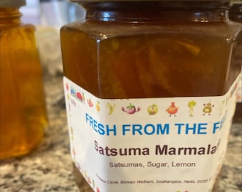 May include: A glass jar of orange marmalade with a white label that reads "FRESH FROM THE FARM Satsuma Marmalade Satsumas, Sugar, Lemon  [address]"