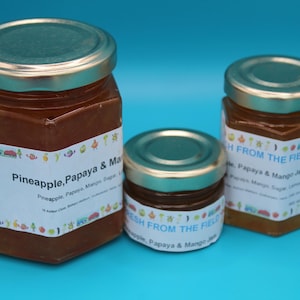 May include: Three glass jars of jam with gold lids. The largest jar is labeled "Pineapple, Papaya & Mango Jam". The two smaller jars are labeled "Fresh From The Field Papaya & Mango Jam".