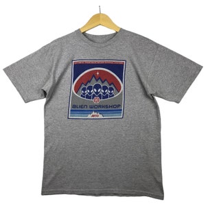 May include: Gray short-sleeved t-shirt with a graphic print of a red, white, and blue alien workshop logo. The logo features a group of aliens in front of a mountain range with a star above them. The text "Alien Workshop" is printed in white below the aliens. The text "ARTO" is printed in white below the text "Alien Workshop".
