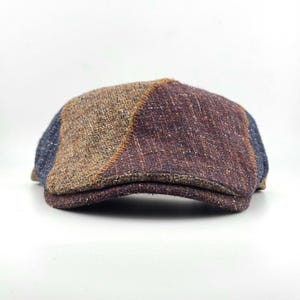 May include: A flat cap featuring a patchwork design with panels of textured fabric in shades of brown, deep purple, and blue. The sections are joined by visible stitching, creating a multi-colored, woven appearance across the crown and brim.