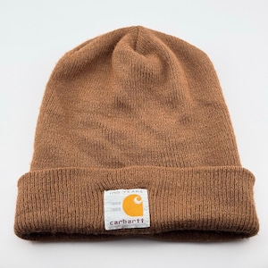 May include: A brown knit beanie with a cuffed brim. A white rectangular patch is sewn onto the center of the cuff, featuring the orange Carhartt logo, the text 100 YEARS, and the years 1889 and 1989 printed in black lettering.