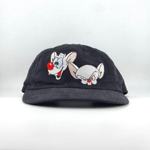 Peut inclure: Casquette de baseball noire avec broderie des personnages de dessins animés Pinky et le Cerveau. Pinky a un nez rouge et un large sourire, tandis que Cerveau a une expression sévère. La casquette est en tissu foncé.
