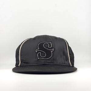 Puede incluir: Gorra de béisbol negra con visera plana. Presenta una gran "S" bordada con contorno blanco en la parte delantera. Ribetes blancos acentúan los laterales, con un diseño sencillo y deportivo.