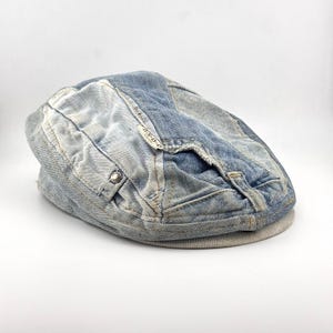 May include: A flat cap constructed from light and medium blue denim fabric panels. The hat features a frayed edge detail, a small metal button, and a white fabric label with the text O.N.O.C. The brim is finished with a textured grey fabric band.